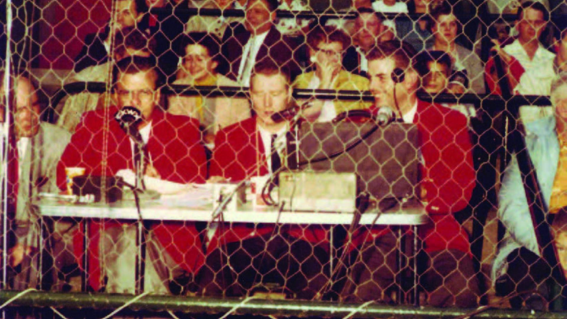 Jim Robson sits between two other broadcasters, calling a game from behind a chain link fence.